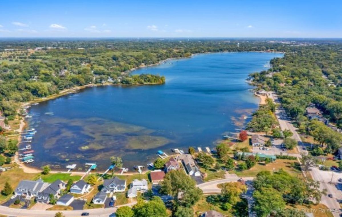 Aerial view of a lake surrounded by houses, docks, and trees, with a road running along the shoreline and a sprawling green landscape in the background under a clear blue sky.