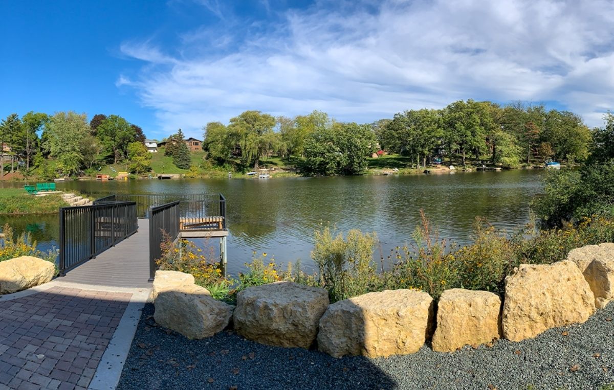 A small wooden dock extends over a calm lake surrounded by trees and greenery under a blue sky with scattered clouds. Large rocks and flowering plants line the shore near the dock.
