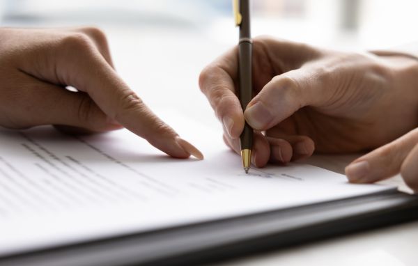 Close-up of one person holding a pen and signing a document while another person points at the paper, indicating where to sign. The focus is on the hands and the paper.