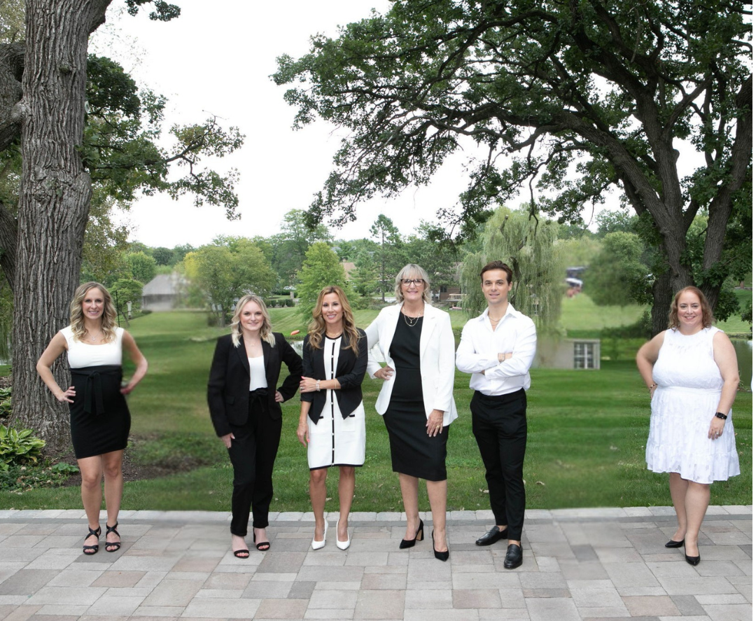 Six people stand outdoors on a paved patio, with green trees and grass in the background. They are dressed formally in black and white attire and are smiling at the camera.