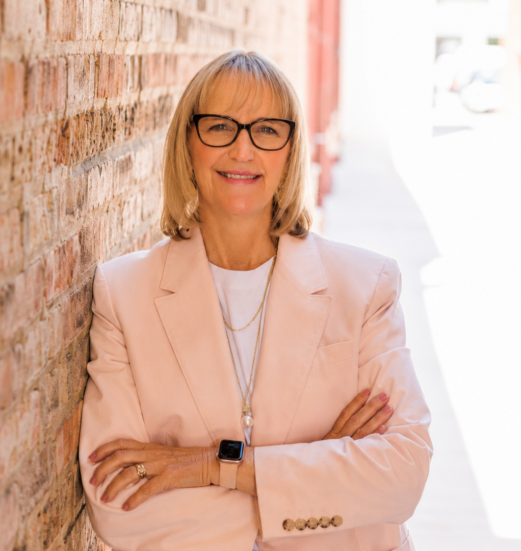 A woman with blonde hair, glasses, and a pink blazer stands outdoors, leaning against a brick wall with her arms crossed, smiling confidently. A woman with blonde hair, glasses, and a pink blazer stands outdoors, leaning against a brick wall with her arms crossed, smiling confidently.