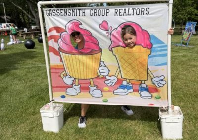 Two kids pose with their faces through cutouts in a cartoon ice cream banner at an outdoor event hosted by Gassensmith Group Realtors. The scene is on grass with trees and games in the background.