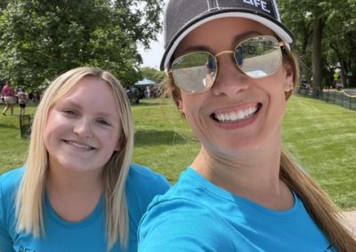 Two women wearing matching blue Real Estate Life shirts smile outdoors in a sunny park. One woman also wears a Real Estate Life cap and sunglasses. Green grass and trees are visible in the background.