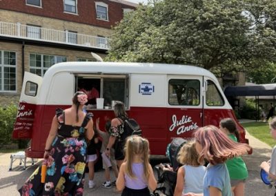 Children gather around a red and white ice cream van labeled Julie Anns while a woman in a floral dress serves treats on a sunny day outside a brick building with trees in the background.