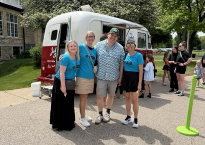 Four adults pose and smile in front of a retro red and white van on a sunny day; others stand in the background near trees and a building.