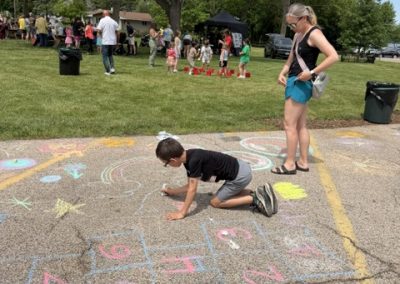 A child draws with chalk on pavement, surrounded by colorful designs and a hopscotch grid, while a woman stands nearby. In the background, people gather on a grassy area under trees.