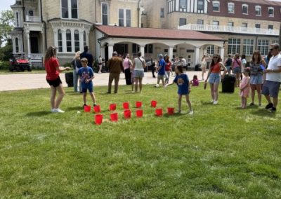 Children and adults gather on a grassy lawn in front of large historic buildings, playing a game with red buckets arranged in rows. Some kids are about to toss balls, while others watch. It’s a bright, partly cloudy day.