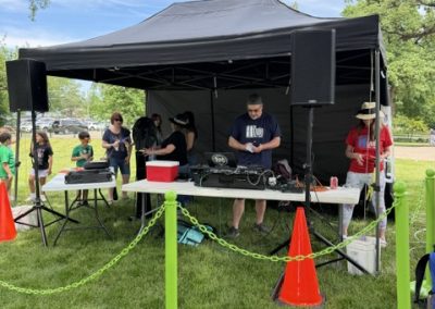 People stand under a black canopy tent at an outdoor event. There are tables with DJ equipment, speakers, and orange cones, with green barriers in front. Children and adults are gathered on the grassy area nearby.
