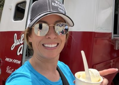 A smiling woman in sunglasses and a black cap holds a cup of ice cream in front of a red-and-white food truck on a sunny day.