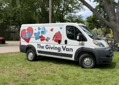 A white van labeled The Giving Van is parked on grass. The van features colorful heart and hand graphics on its side. Trees and houses are visible in the background.