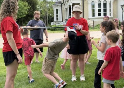 A group of children and adults play limbo outside on grass near a large building. Two women hold the limbo stick while a boy bends under it. Others wait and watch, enjoying the outdoor activity on a sunny day.