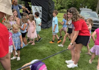 Children and adults play limbo outside on grass. A girl bends backward under a rope, while others wait in line. Two adults in red shirts hold the rope. There are tents and trees in the background.