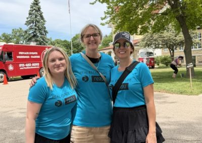 Three women wearing matching blue Real Estate shirts and black skirts or shorts stand smiling outdoors on a sunny day, with green trees and a red truck in the background.