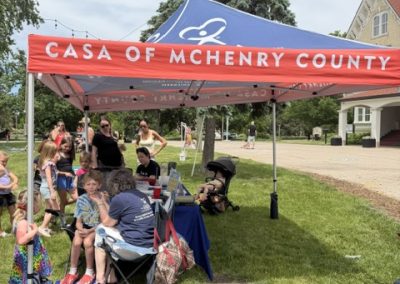 A red tent labeled Casa of McHenry County shelters adults and children at an outdoor event. Kids sit and play while adults stand nearby. Trees and a large building are in the background under a partly cloudy sky.