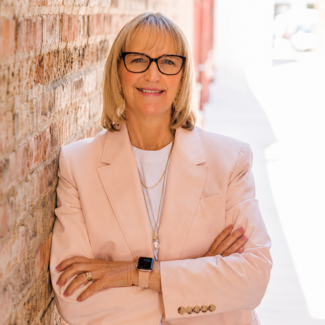 A woman with blonde hair and glasses stands against a brick wall, smiling with her arms crossed. She is wearing a light pink blazer, layered necklaces, and a smartwatch. The background is brightly lit.