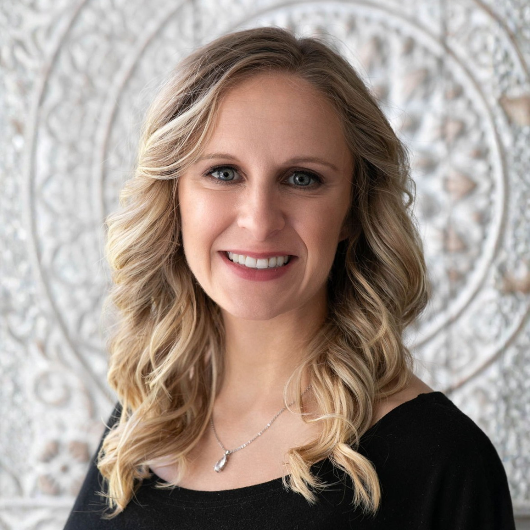 A woman with wavy blonde hair, wearing a black top and a necklace, smiles in front of a textured, decorative white background.